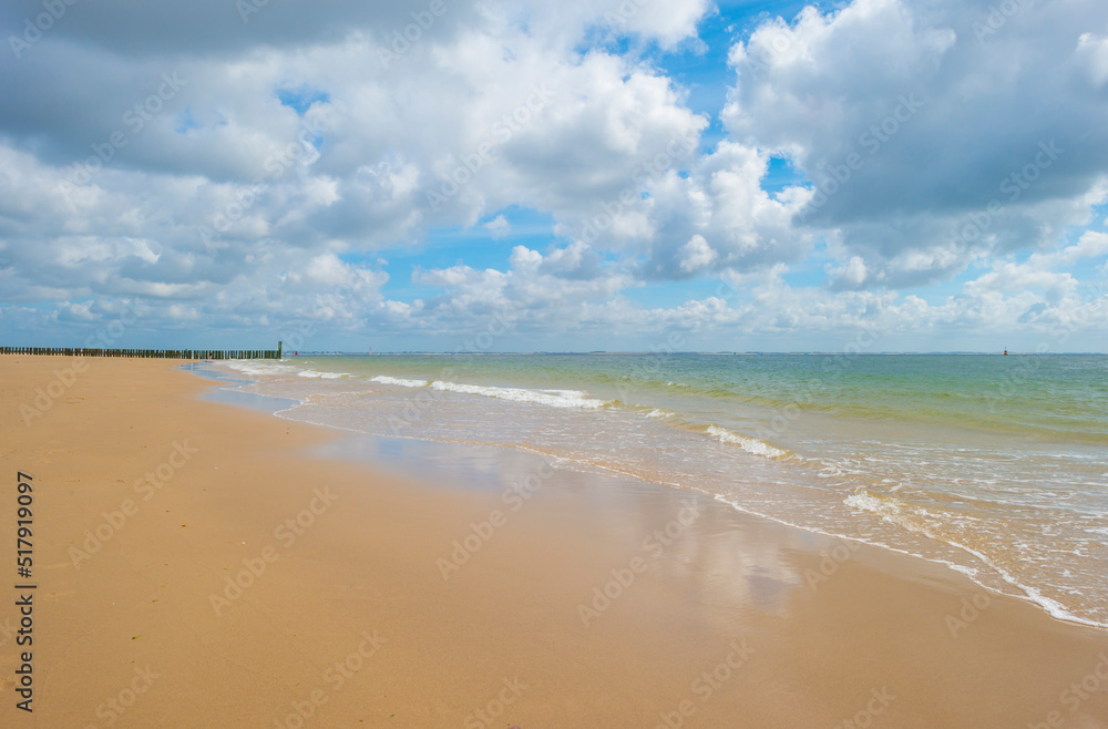 Sunlit waves and a wooden breakwater on the yellow sand of a sunny beach along a sea under a blue cloudy sky in summer, Walcheren, Zeeland, the Netherlands, July, 2022