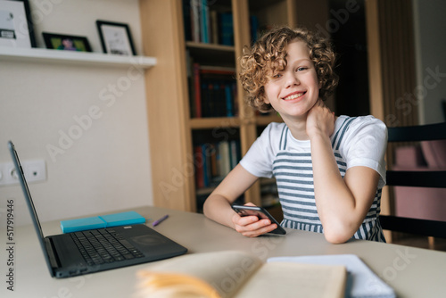 Portrait of smiling adorable preteen girl looking at camera sitting at desk, holding smartphone in hands, studying online, writing notes, using laptop, watching webinar, listening to lecture, at home.