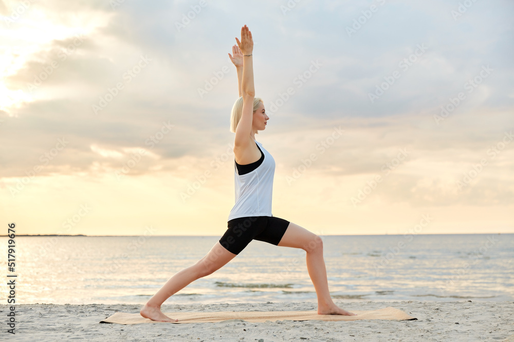 fitness, sport, and healthy lifestyle concept - happy woman doing yoga warrior pose on beach over sunset