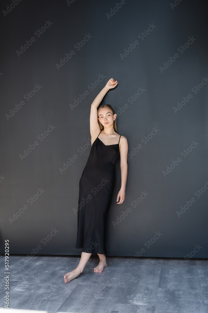 full length of barefoot woman in black strap dress posing with raised hand near dark wall.
