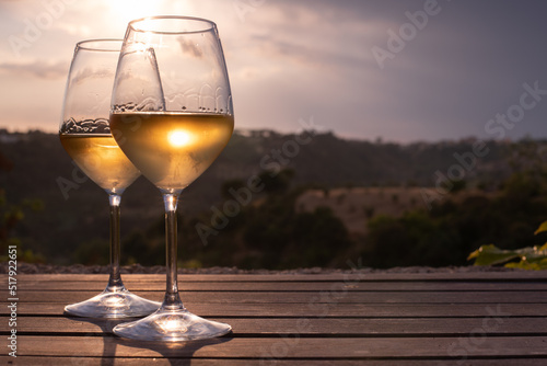 Two elegant glasses of white wine on the right, and a countryside view in the background, at sunset