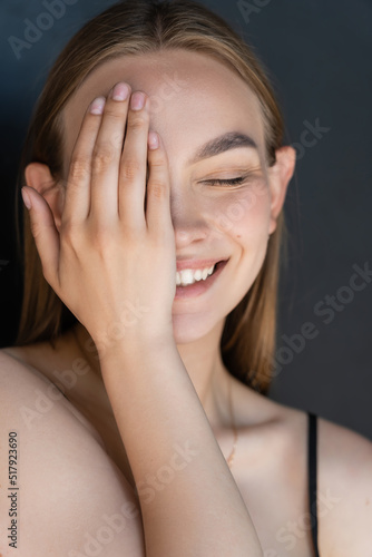 portrait of smiling woman with natural makeup obscuring face with hand isolated on black.