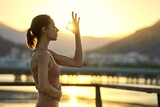 Young sporty woman practicing yoga and meditation outdoor during sunset against mountain range. Girl makes mudra gesture with sunlight