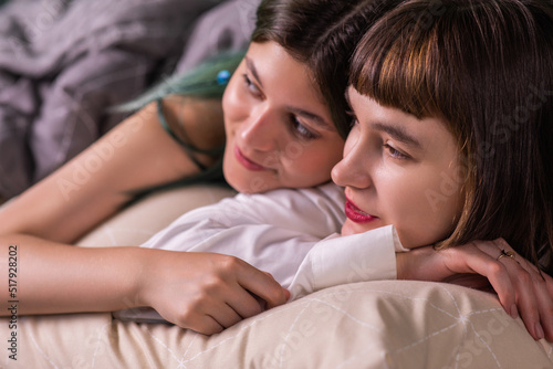 Amazing ladies young looking lesbian couple posing in front of the camera while laying on the bed together hugging each other with love and looking straight