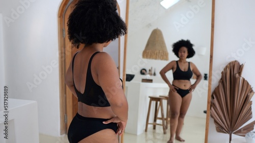 A plump black woman in front of a mirror looks at her body. A plus size model in front of a mirror in her lingerie feels body-positive and doesn't consider herself fat. Self acceptance concept.