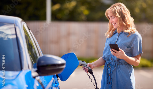 Принти на полотні Woman Charging Electric Car With Cable Using App On Phone To Monitor Battery Lev