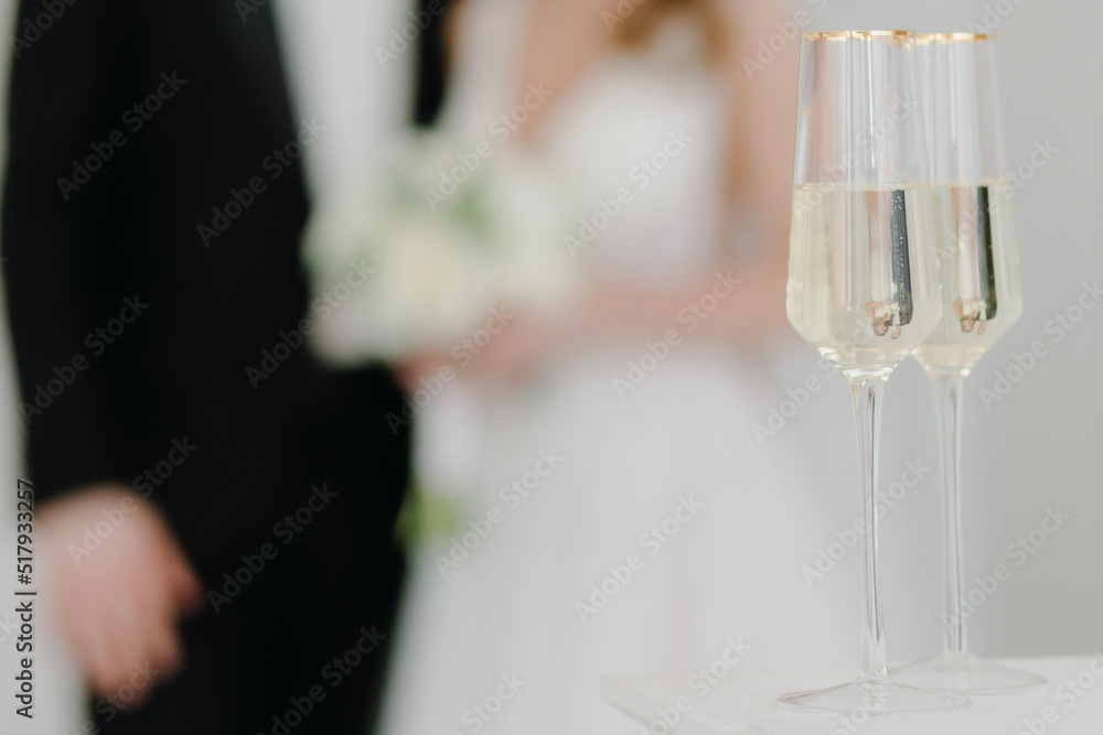 Two glasses of champagne stand on the table against the background of newlyweds in love at the wedding ceremony. Two glasses of champagne stand on the table against the background of newlyweds in love at the wedding ceremony.
