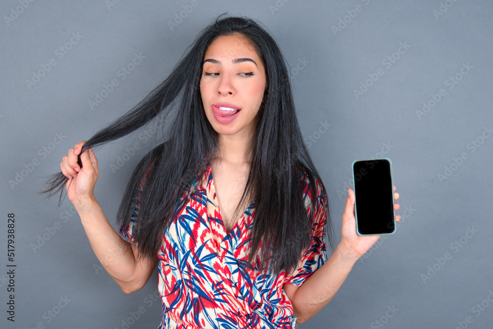 Photo of nice pretty Young latin woman wearing colorful dress over grey backgrogund demonstrate phone screen hold hair tails Photo of nice pretty Young latin woman wearing colorful dress over grey backgrogund demonstrate phone screen hold hair tails