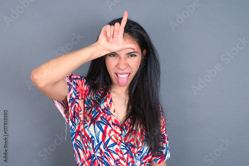 Funny Young latin woman wearing colorful dress over grey backgrogund makes loser gesture mocking at someone sticks out tongue making grimace face. photo