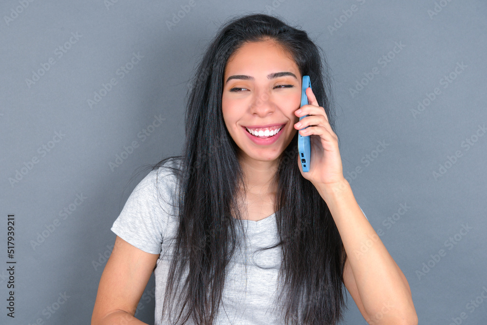 Funny young beautiful brunette woman wearing grey t-shirt over grey background laughs happily, has phone conversation, being amused by friend, closes eyes. Funny young beautiful brunette woman wearing grey t-shirt over grey background laughs happily, has phone conversation, being amused by friend, closes eyes.