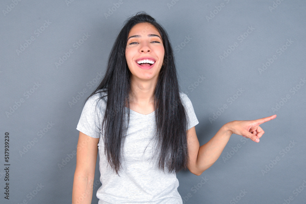 young beautiful brunette woman wearing white t-shirt over grey background laughs happily points away on blank space demonstrates shopping discount offer, excited by good news or unexpected sale. young beautiful brunette woman wearing white t-shirt over grey background laughs happily points away on blank space demonstrates shopping discount offer, excited by good news or unexpected sale.