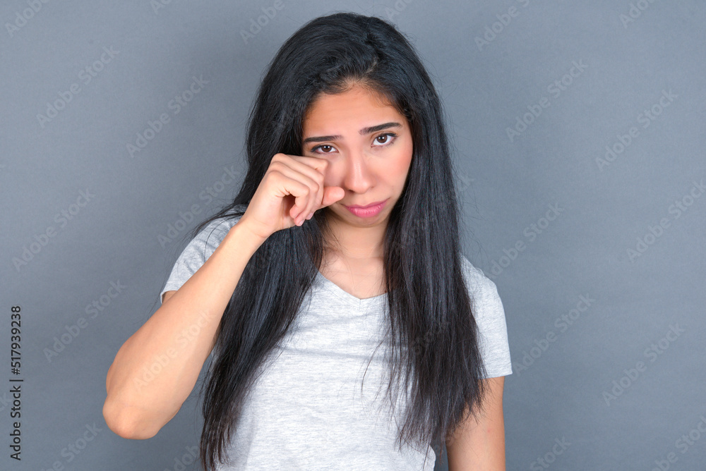 Disappointed dejected young beautiful brunette woman wearing white t-shirt over grey background wipes tears stands stressed with gloomy expression. Negative emotion Disappointed dejected young beautiful brunette woman wearing white t-shirt over grey background wipes tears stands stressed with gloomy expression. Negative emotion