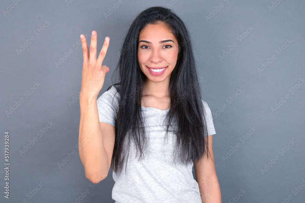 young beautiful brunette woman wearing white t-shirt over grey background smiling and looking friendly, showing number three or third with hand forward, counting down young beautiful brunette woman wearing white t-shirt over grey background smiling and looking friendly, showing number three or third with hand forward, counting down
