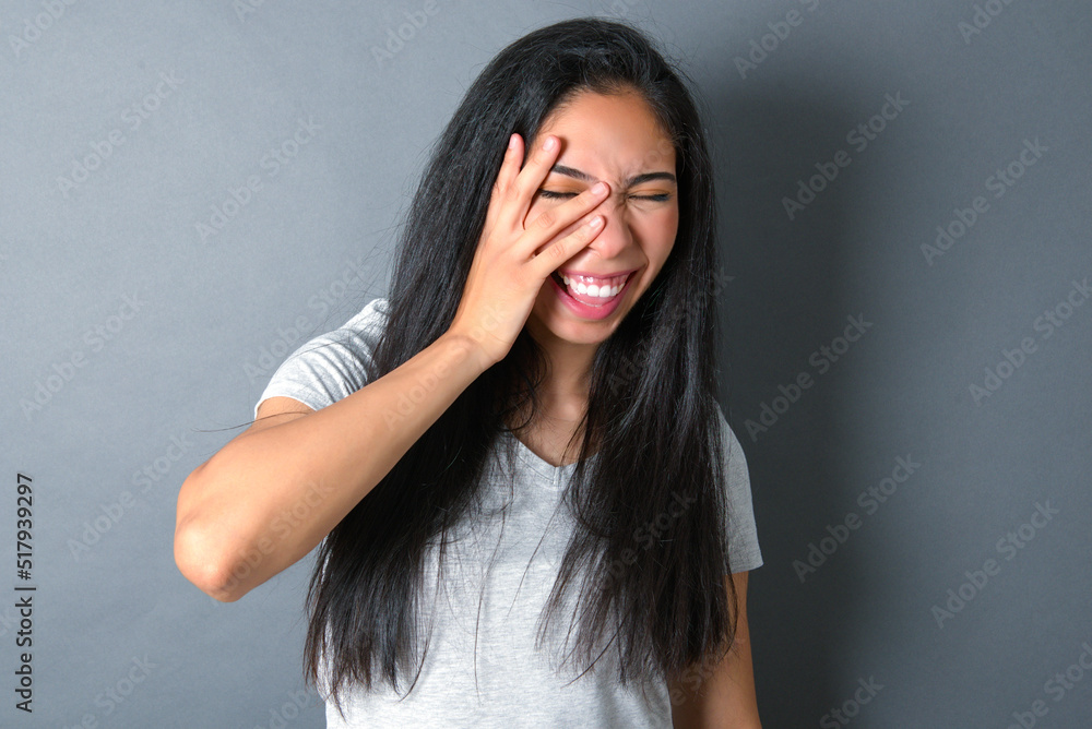 young beautiful brunette woman wearing white t-shirt over grey background makes face palm and smiles broadly, giggles positively hears funny joke poses young beautiful brunette woman wearing white t-shirt over grey background makes face palm and smiles broadly, giggles positively hears funny joke poses