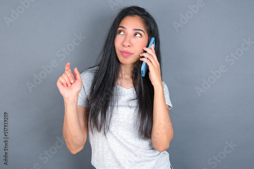 young beautiful brunette woman wearing grey t-shirt over grey background speaks on mobile phone spends free time indoors calls to friend.