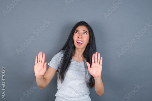 young beautiful brunette woman wearing white t-shirt over grey background keeps palms forward and looks with fright above on ceiling tries to defense herself from invisible danger opens mouth.