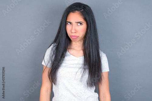 Dissatisfied young beautiful brunette woman wearing white t-shirt over grey background purses lips and has unhappy expression looks away stands offended. Depressed frustrated model.