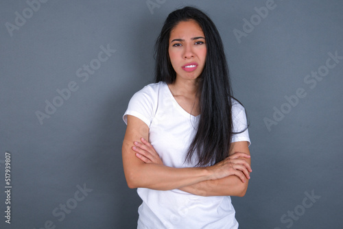 young beautiful brunette woman wearing white t-shirt over grey background frowning his face in displeasure, keeping arms folded, waiting for an explanation.