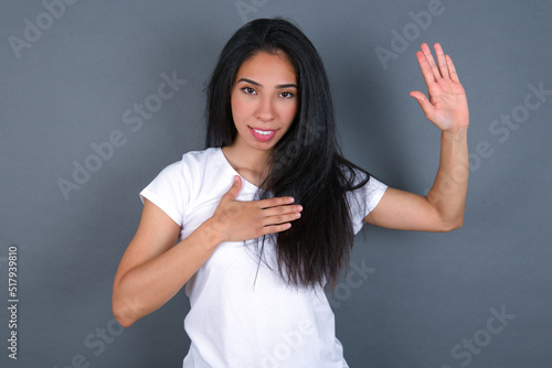 I swear, promise you not regret. Portrait of sincere young beautiful brunette woman wearing white t-shirt over raising one arm and hold hand on heart as give oath, telling truth, want you to believe.