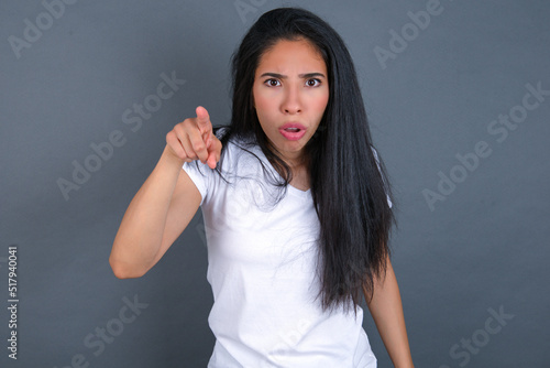 Shocked young beautiful brunette woman wearing white t-shirt over grey background points front with index finger at camera and. Surprise and advertisement concept.