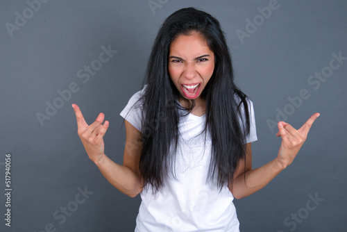 Born to rock this world. Joyful young beautiful brunette woman wearing white t-shirt over grey background screaming out loud and showing with raised arms horns or rock gesture.