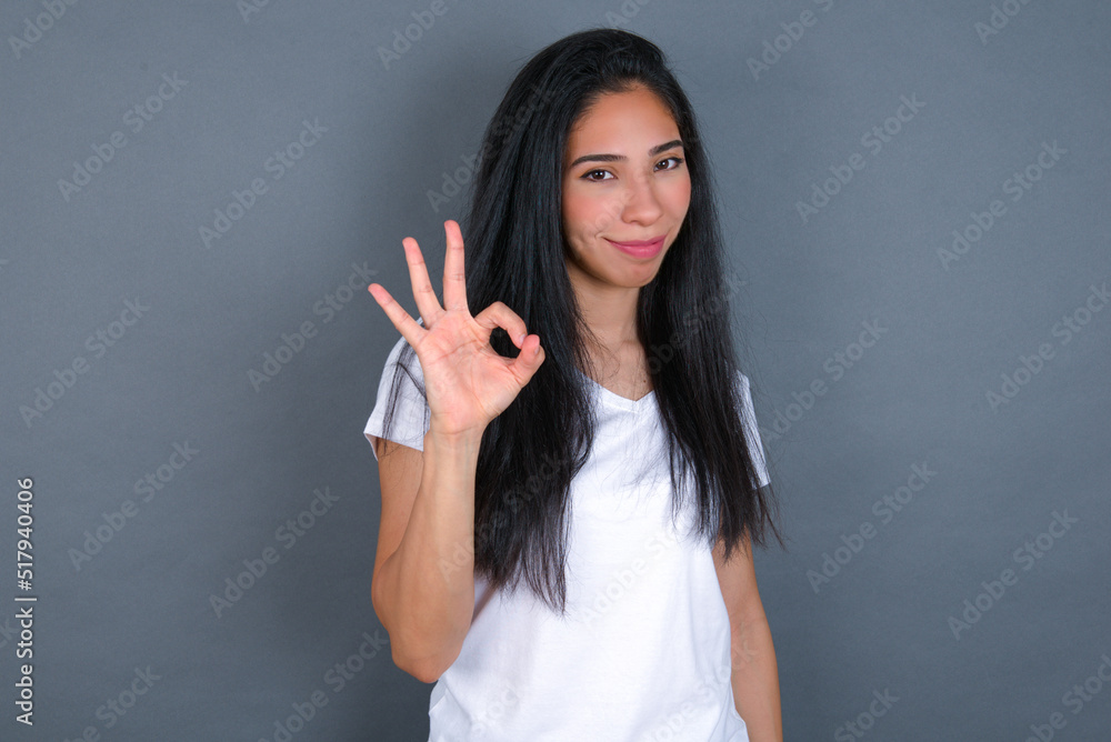 Glad attractive young beautiful brunette woman wearing white t-shirt over grey background shows ok sign with hand as expresses approval, has cheerful expression, being optimistic. Glad attractive young beautiful brunette woman wearing white t-shirt over grey background shows ok sign with hand as expresses approval, has cheerful expression, being optimistic.