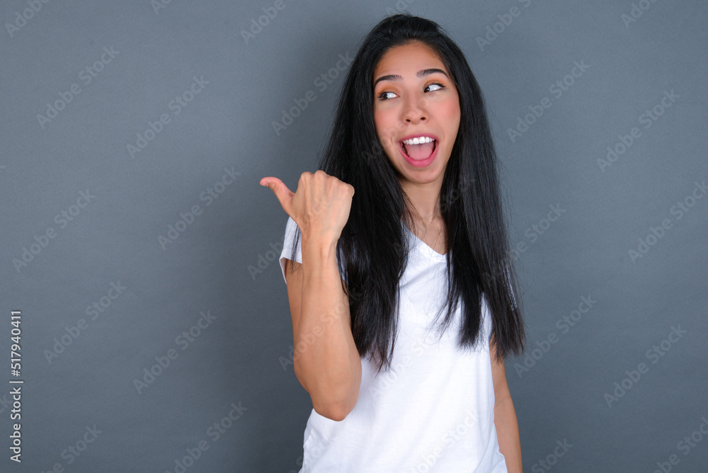 Stupefied young beautiful brunette woman wearing white t-shirt over grey background with surprised expression, opens eyes and mouth widely, points aside with thumb, shows something strange. Stupefied young beautiful brunette woman wearing white t-shirt over grey background with surprised expression, opens eyes and mouth widely, points aside with thumb, shows something strange.