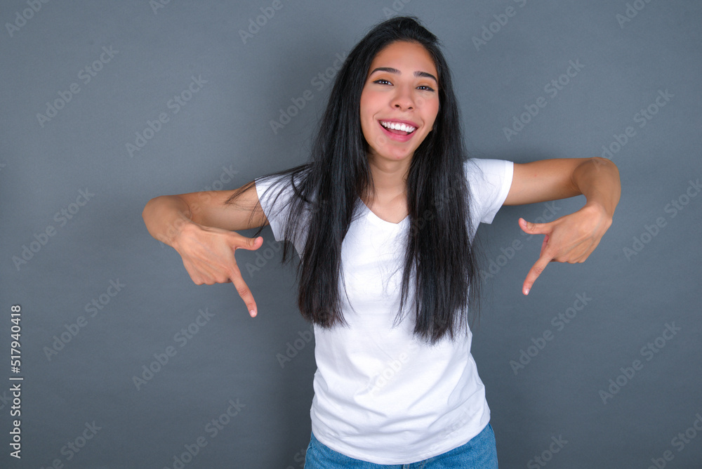 young beautiful brunette woman wearing white t-shirt over grey background with positive expression, points down with both index fingers, has broad interested smile. Look there, please. young beautiful brunette woman wearing white t-shirt over grey background with positive expression, points down with both index fingers, has broad interested smile. Look there, please.