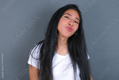 young beautiful brunette woman wearing white t-shirt over grey background, keeps lips as going to kiss someone, has glad expression, grimace face. Standing indoors. Beauty concept.