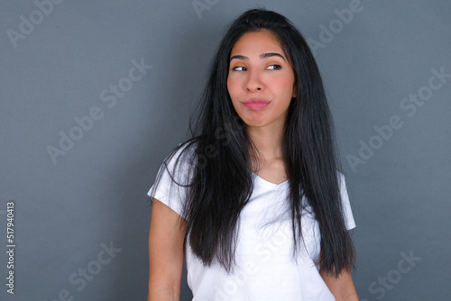 young beautiful brunette woman wearing white t-shirt over grey background, looks pensively aside, plans actions after university, imagines what to do Thinks over about new project.