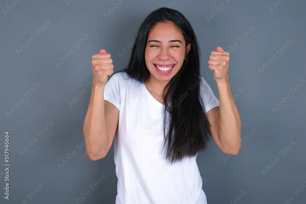young beautiful brunette woman wearing white t-shirt over grey background rejoicing his success and victory clenching fists with joy being happy to achieve aim and goals. Positive emotions, feelings. young beautiful brunette woman wearing white t-shirt over grey background rejoicing his success and victory clenching fists with joy being happy to achieve aim and goals. Positive emotions, feelings.