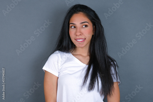 young beautiful brunette woman wearing white t-shirt over grey background with thoughtful expression, looks away keeps hands down bitting his lip thinks about something pleasant.