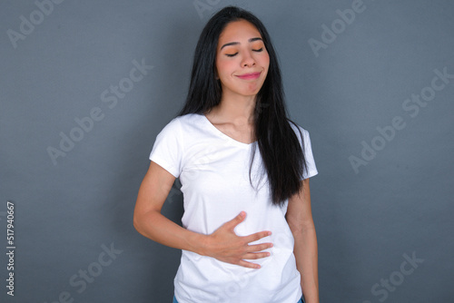 Satisfied smiling young beautiful brunette woman wearing white t-shirt , keeps hands on belly, being in good mood after eating delicious supper, demonstrates she is full. Pleasant feeling in stomach.