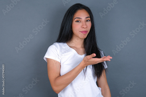 young beautiful brunette woman wearing white t-shirt over grey background says: wow how exciting it is, has amazed expression, shows something on blank space with palm. Advertisement concept.