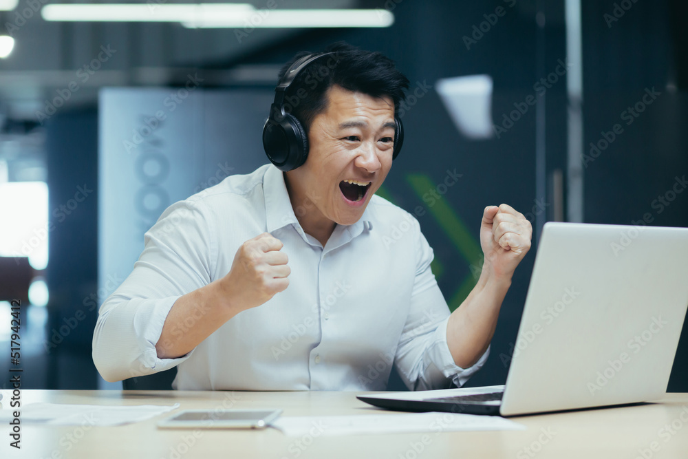 Happy Asian businessman watching sports match online, man in office looking at laptop screen cheerfully cheering Happy Asian businessman watching sports match online, man in office looking at laptop screen cheerfully cheering