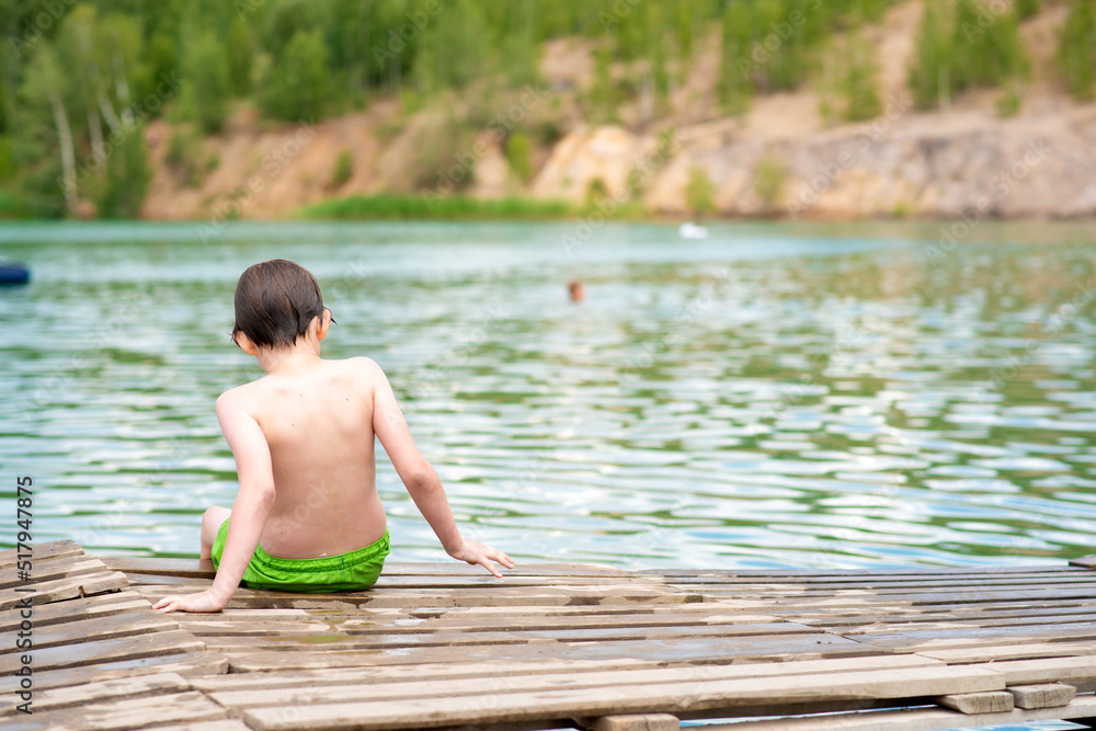a little boy sits on a wooden bridge after swimming in the lake. wet child sits on the shore of the lake. a little boy sits on a wooden bridge after swimming in the lake. wet child sits on the shore of the lake.