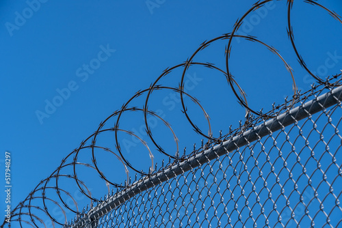 Fence with razor wire with blue sky in the background is shown. 
Razor wire is made up of high tensile core wire and a punched steel tape with sharp barbs at close intervals uniformly.
 photo