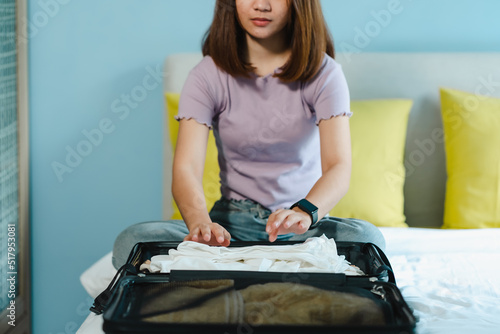 A teenage Asian woman folds up her shirt and pants in a suitcase on her bed to prepare for a weekend trip. Pack your luggage and check forgot anything before going on trip. © Ekkasit A Siam