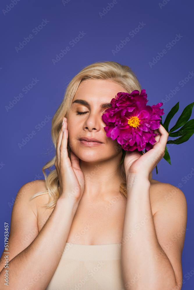 blonde woman with bare shoulders covering eye with blooming flower isolated on purple. blonde woman with bare shoulders covering eye with blooming flower isolated on purple.