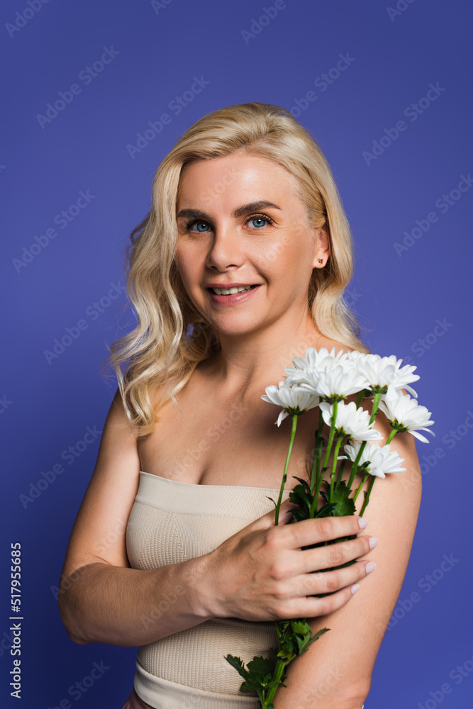 cheerful woman with bare shoulders looking at camera while holding flowers isolated on violet. cheerful woman with bare shoulders looking at camera while holding flowers isolated on violet.