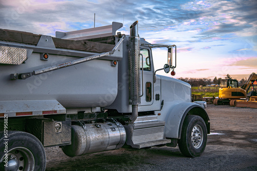 Construction truck. A gray commercial construction truck in the foreground. An excavator is in the background. Cloudy sky blurry.