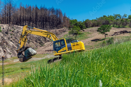 Mountainous terrain with an excavator in the foreground. A large stone in the bucket of an excavator. Clearing the square after a natural disaster. Burnt trees.