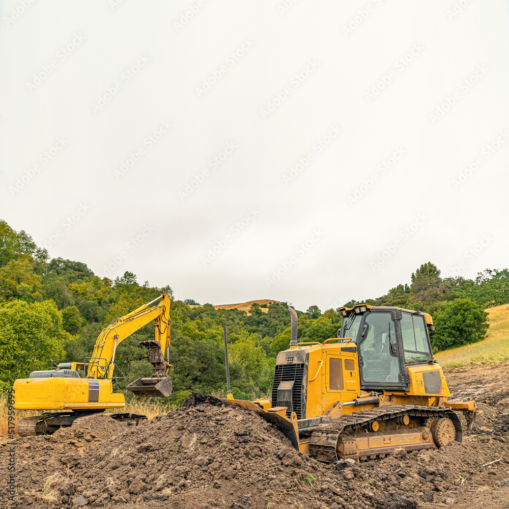 View of a yellow bulldozer and an excavator on a plot of land. Heavy equipment clears the area. Forest slopes background.. View of a yellow bulldozer and an excavator on a plot of land. Heavy equipment clears the area. Forest slopes background..