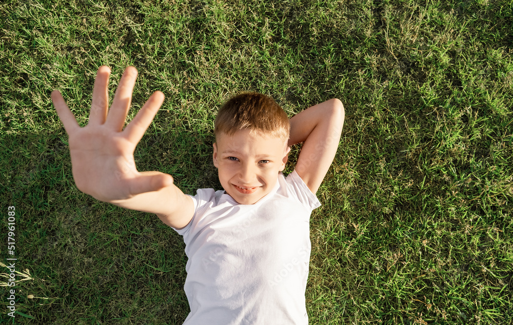 boy lies on grass with smile and puts out his hand. Portrait of schoolboy in white t-shirt boy lies on grass with smile and puts out his hand. Portrait of schoolboy in white t-shirt