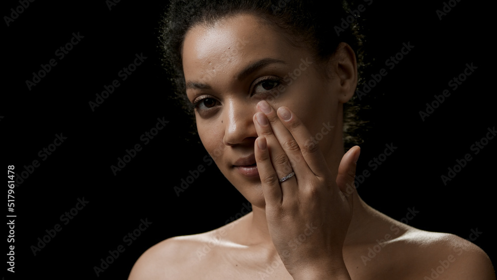 Pretty slim African American young woman with dark curly hair touches her cheek looking at the camera on black background | Face moisturizing concept Pretty slim African American young woman with dark curly hair touches her cheek looking at the camera on black background | Face moisturizing concept