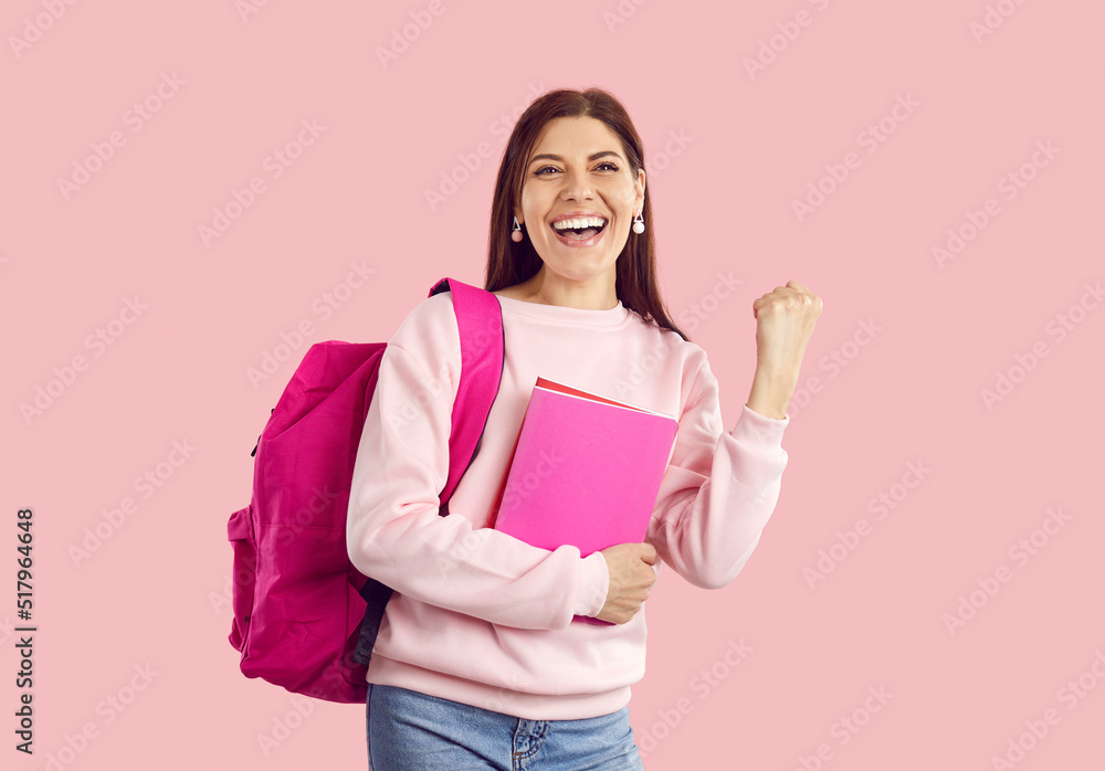 Happy excited female student with backpack and notebooks celebrates exams on pink background. Portrait of young woman with backpack, textbooks and in casual clothes clenching her fists shouting yes. Happy excited female student with backpack and notebooks celebrates exams on pink background. Portrait of young woman with backpack, textbooks and in casual clothes clenching her fists shouting yes.