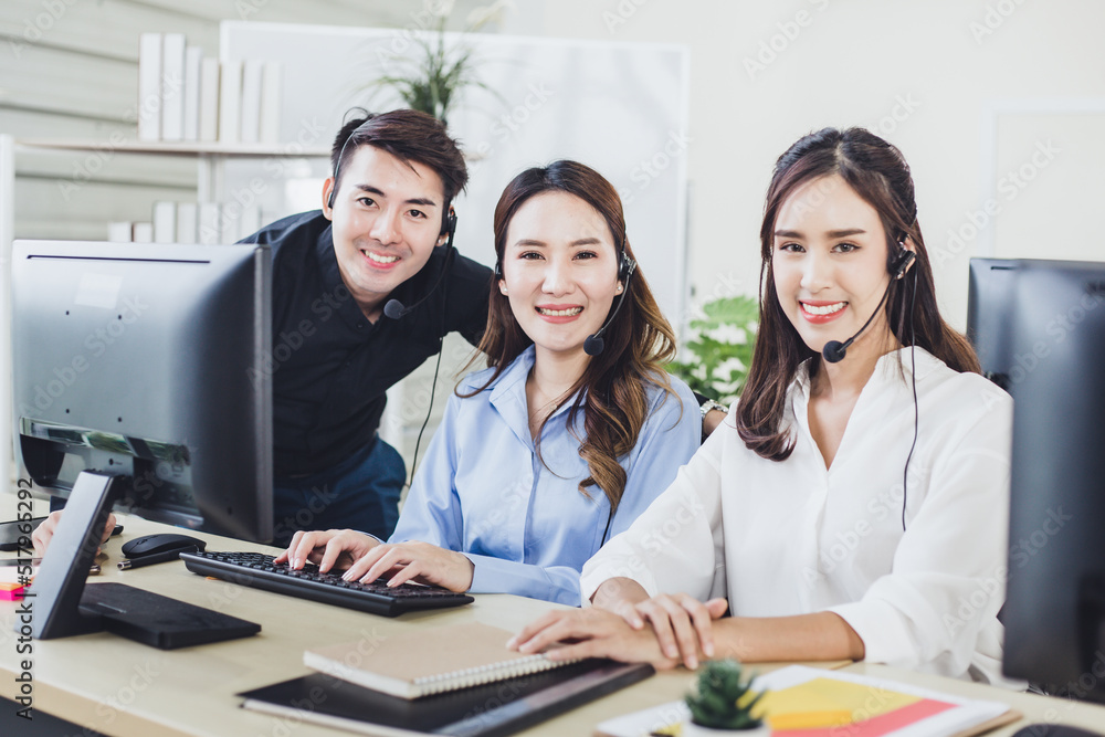 Successful Asian customer service team looking at the camera while working in call center. Group of young smiling operator with headset in modern office. Successful Asian customer service team looking at the camera while working in call center. Group of young smiling operator with headset in modern office.
