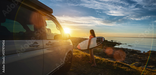 Surfer girl walking near her mini van and looking on the ocean at summer sunset with a surfboard on her side
