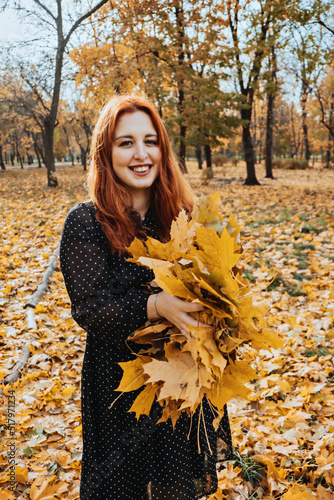 Happy red-haired woman holding yellow maple autumn leaves in fall park.
