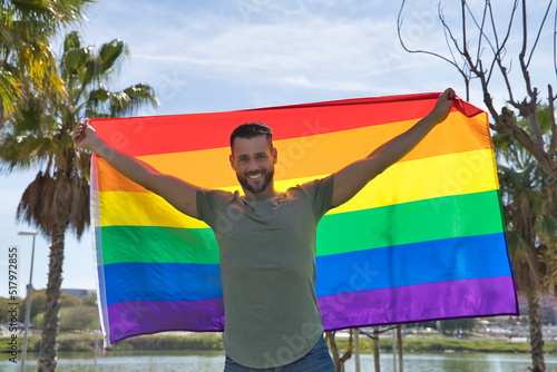 Young and handsome gay man  with a beard and green shirt  with blue eyes  perfect smile and holding a gay pride flag  smiling. Concept of gay pride  homosexual  lgtbi  pride day.
